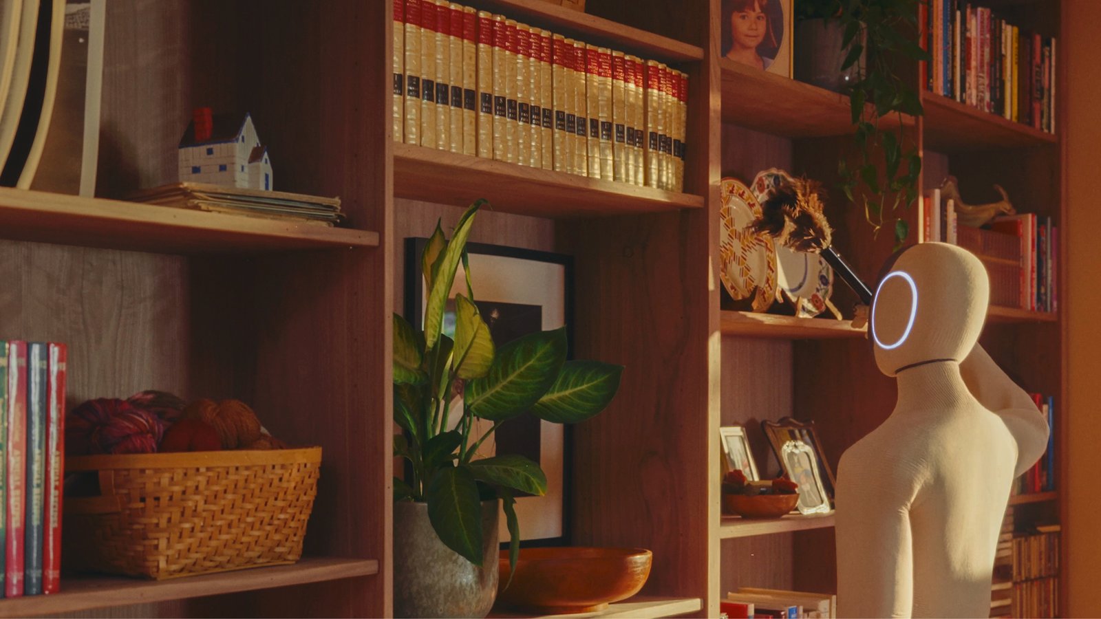 Cream-coloured NEO humanoid robot dusting a wooden bookshelf with a feather duster