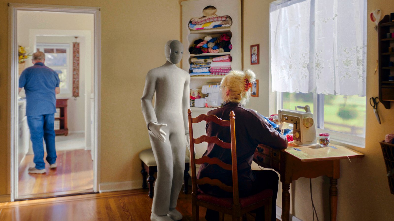 Grey NEO robot standing in a sewing room beside a woman at a sewing machine while a man walks through the doorway